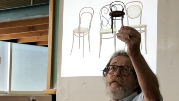 A man, Stergios Delialis, holding up a miniature of a chair while an image of three chairs is projected behind him.