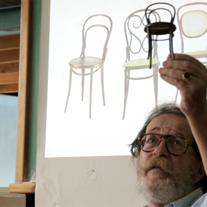 A man, Stergios Delialis, holding up a miniature of a chair while an image of three chairs is projected behind him.