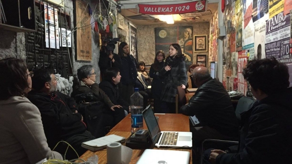 A woman addressing a room of people, posters on the walls and a flag behind her.