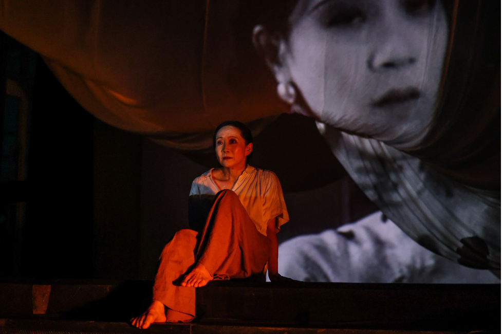 Woman lit in red lighting in front of a screen with a woman's face projected onto it