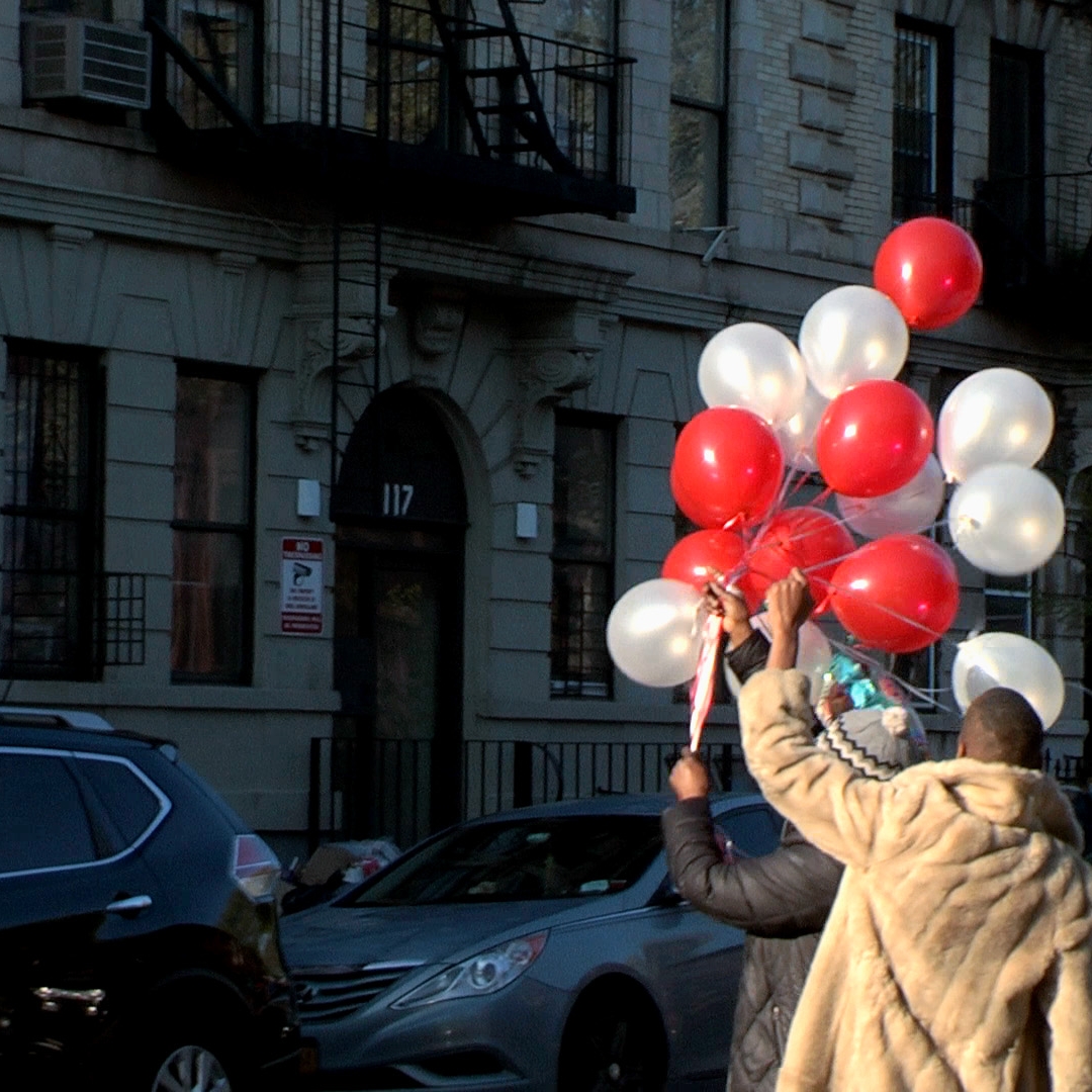 Two people holding red and white balloons in the air on a street corner with cars in the background