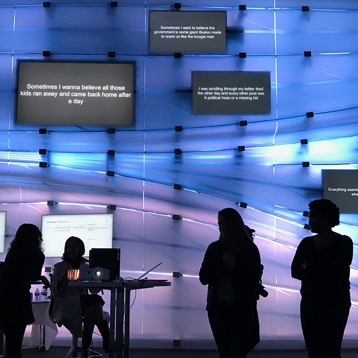 Figures silhouetted against a wall that is blue and purple, with monitors showing news poetry