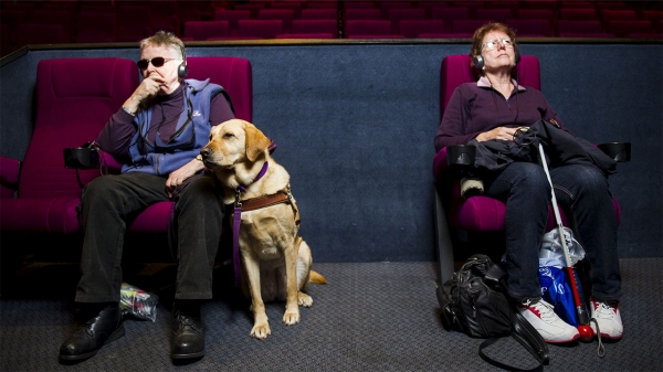 Image Description:Two middle-aged women seated in a movie theater, one with a guide dog and the other with a white cane, sit with head phones on, absorbed in audio description. Photo: Rohan Thomson, courtesy of The Canberra Times