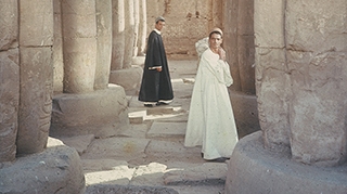 Two figures standing between stone pillars, one in the background wearing black, the one in the foreground wearing white