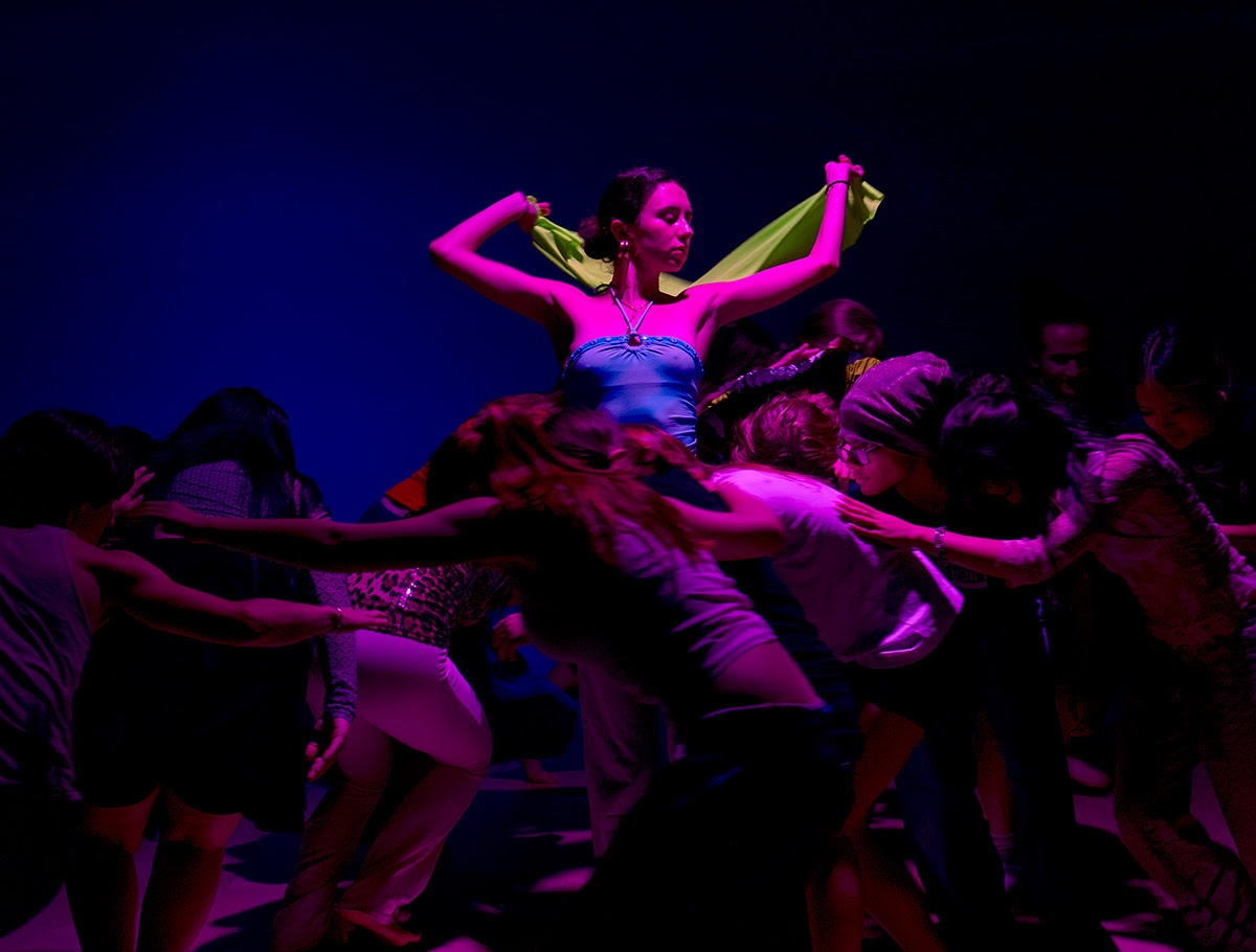 Student in a blue shirt holding a green scarf bathed in pink light, other students crouch and form a circle around her