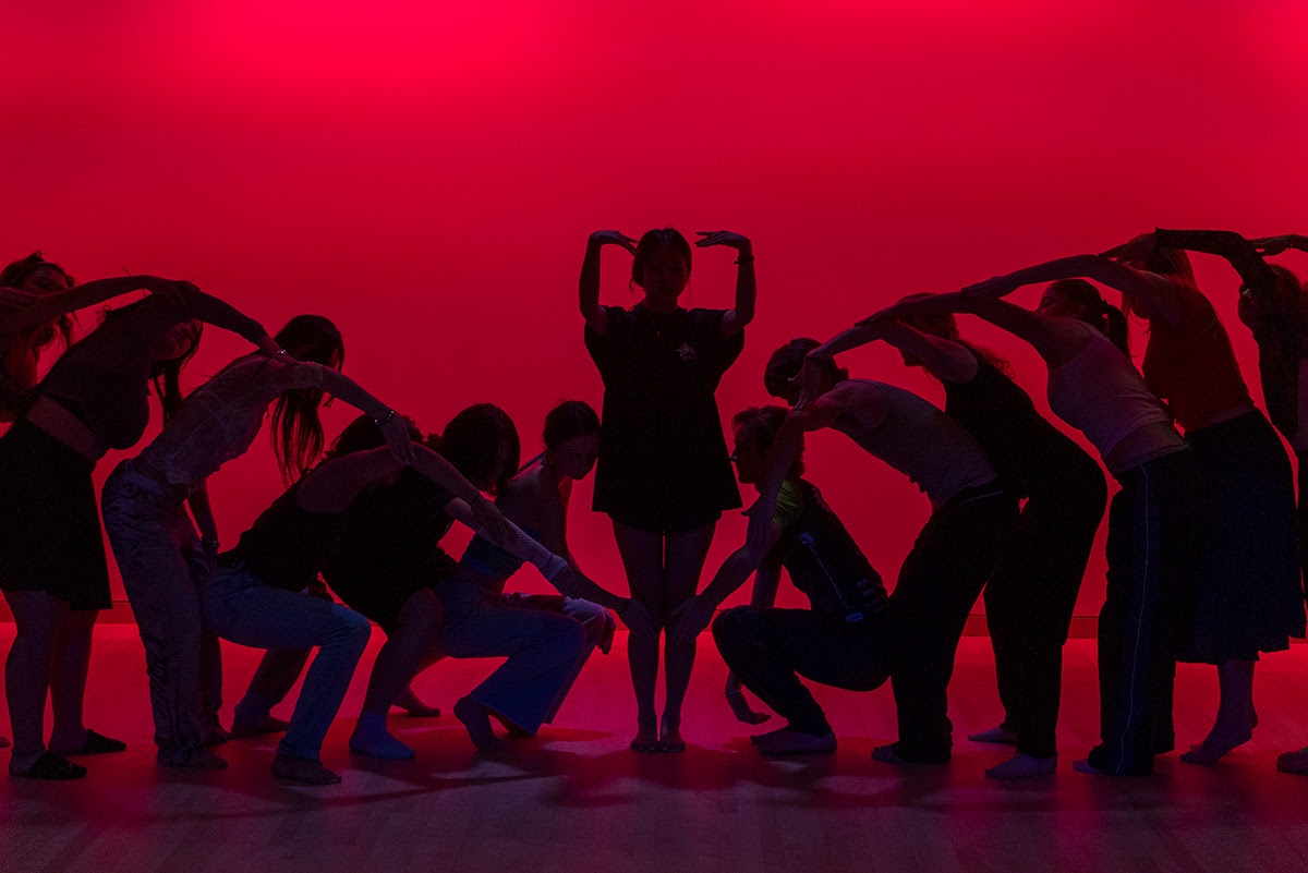 Outlines of students posed symmetrically, arms forming an arch, with one student standing in the center, bathed in red light