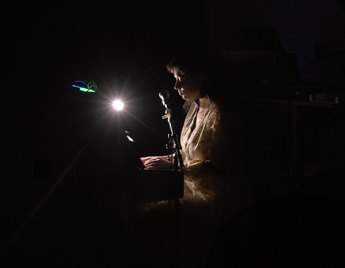 Anatolia sings and plays piano, lit by a single bulb near her face