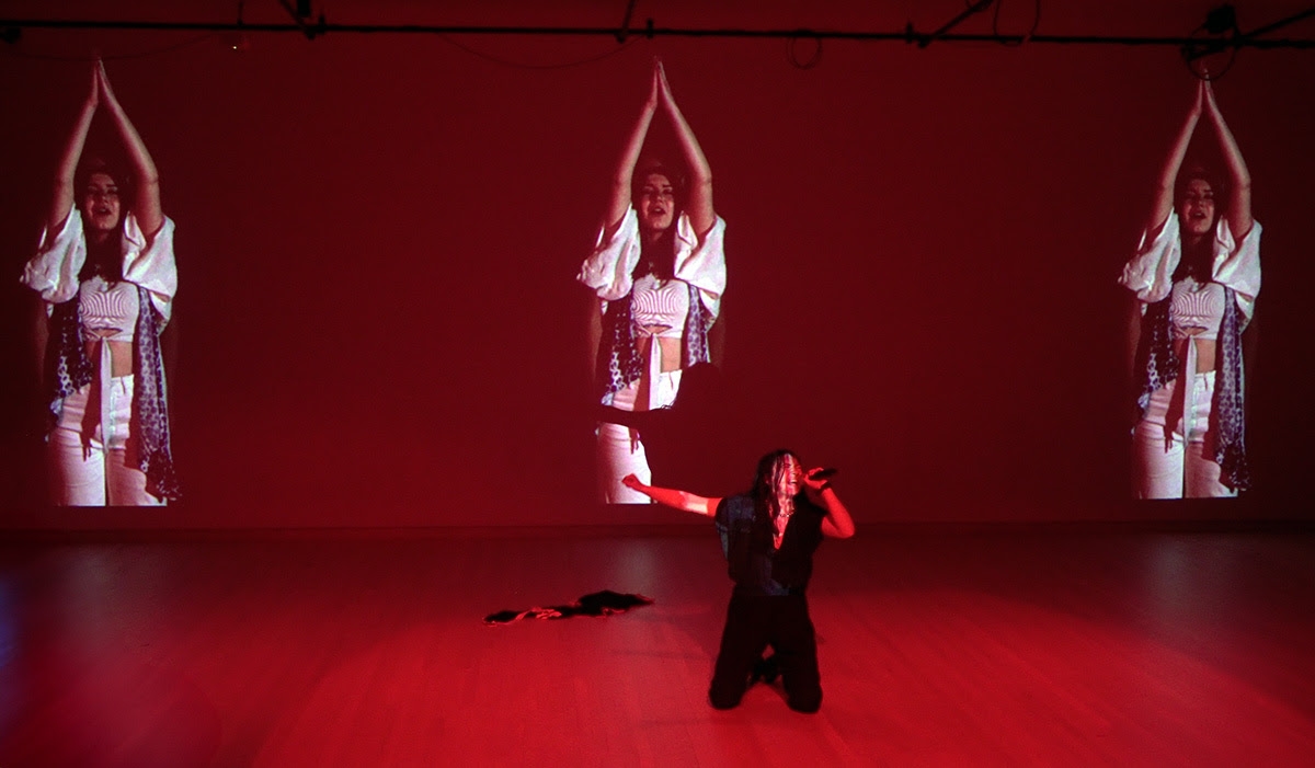 Student kneels and sings into microphone bathed in red light, 3 duplicate images of her are projected onto the wall behind her