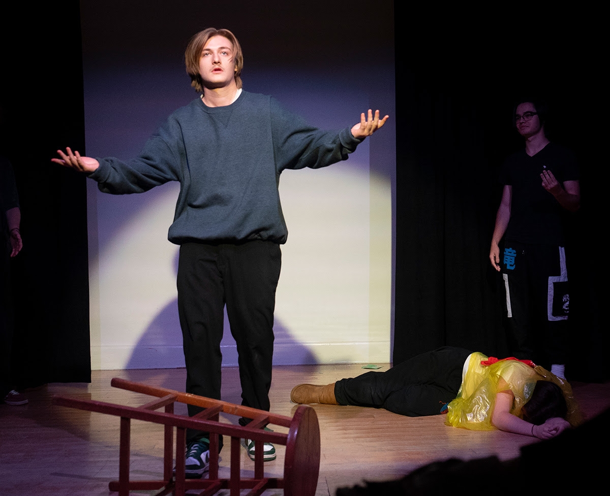 Student in blue sweater poses with arms wide under a spotlight, a stool knocked over in front of him