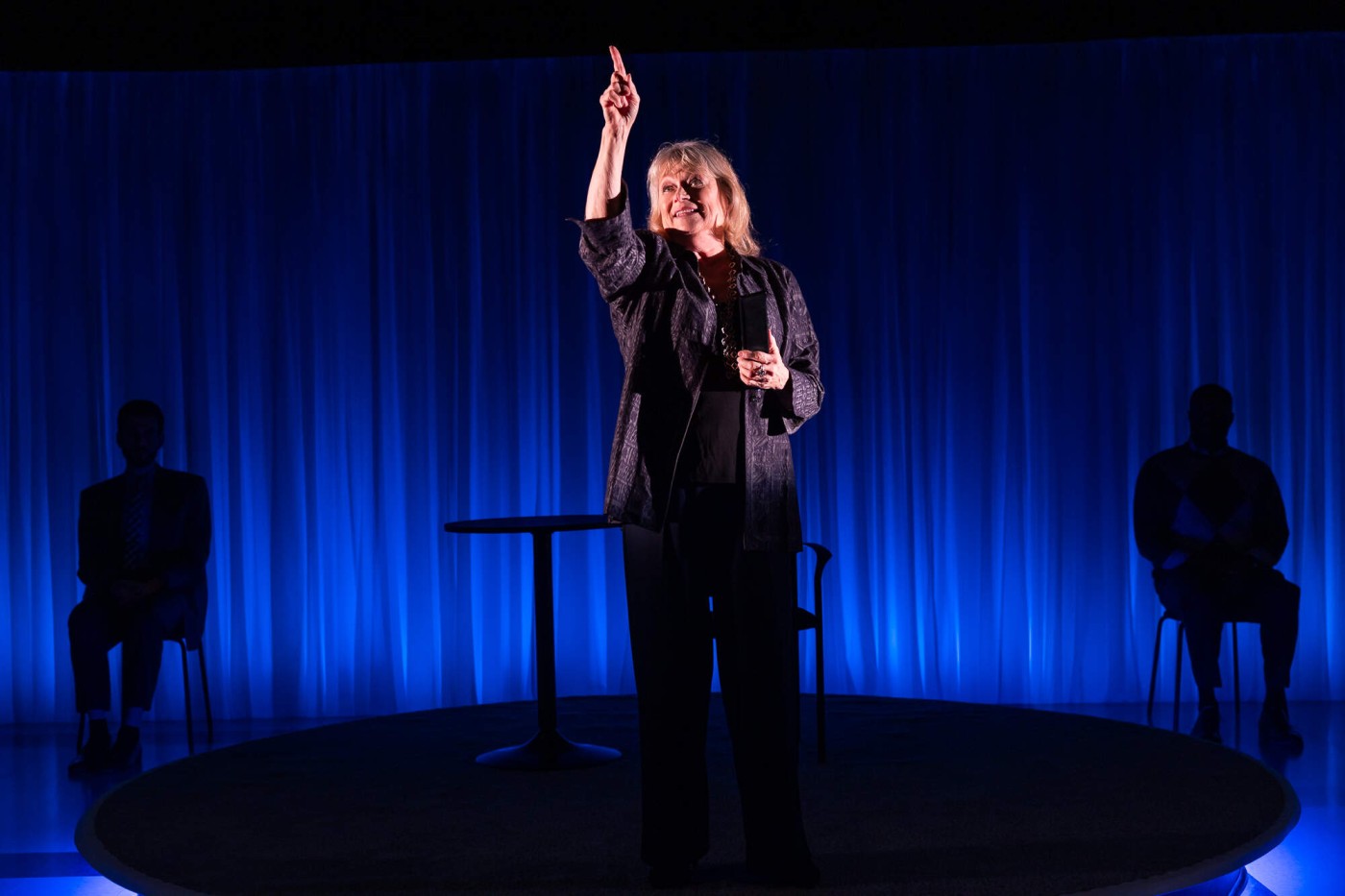 Karen MacDonald stands under spotlight centerstage against a blue background, with one hand pointed upward