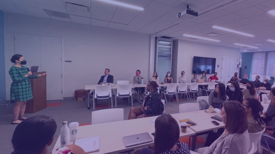 A facilitator stands to the left of the image. A classroom full of diverse individuals listens intently.