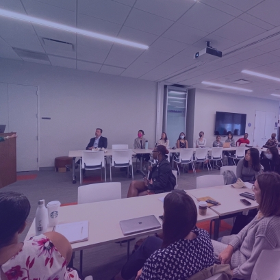 A facilitator stands to the left of the image. A classroom full of diverse individuals listens intently.