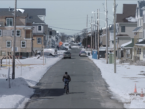 Film still from A New Year. Image looking down the street with boy on bike. 