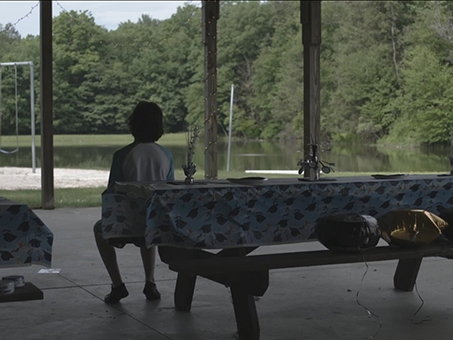 Film Still from Enchantment Falls. Image of young man sitting at picnic table looking out into the distance. 