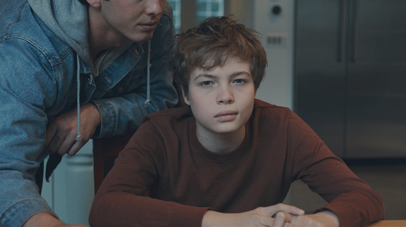 A young boy is centered, sitting at a table, with another young man leaning over him.