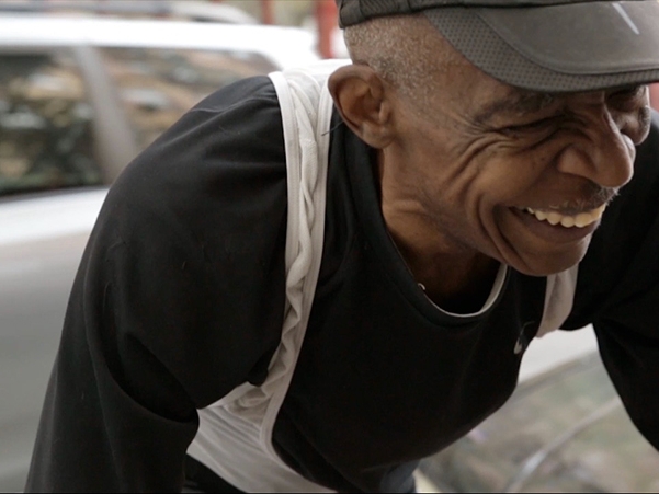 Film still from Who is Alex Harsley? Image of man smiling with cap on. 