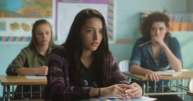 Three young girls in a classroom, sitting at separate desks.