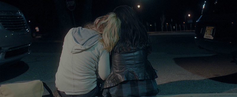 Two women in an embrace while sitting on a street curb.