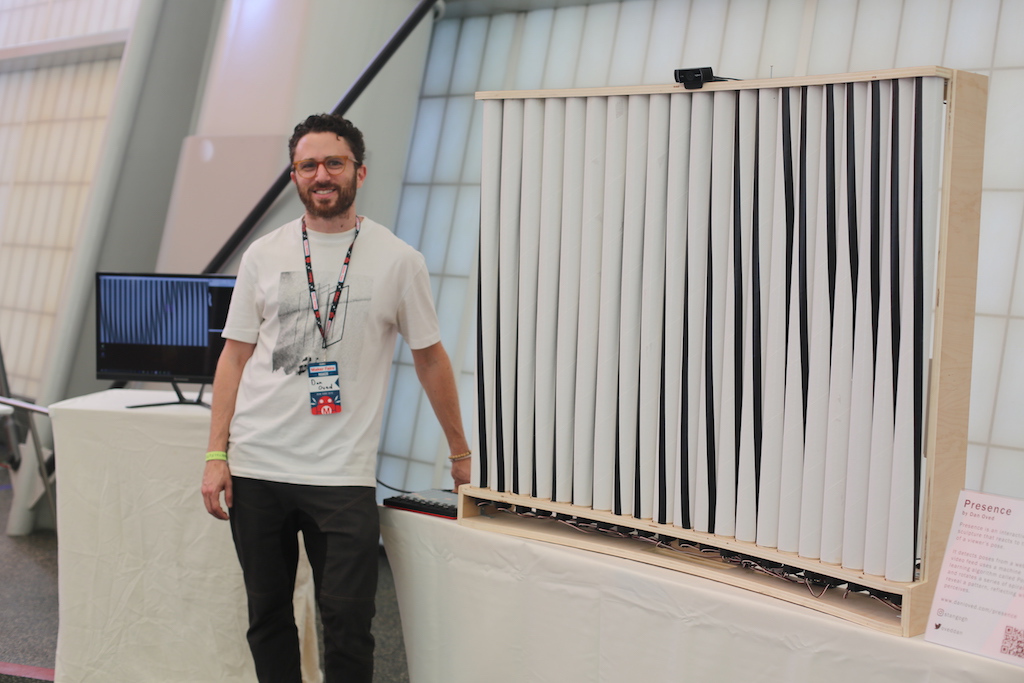 Student stands with project of long stripes of plastic attached to wooden frame