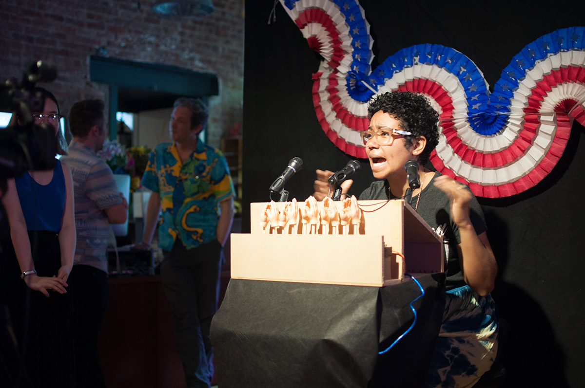 a woman talking into a microphone with a row of mini hands applauding and an american flag banner behind her