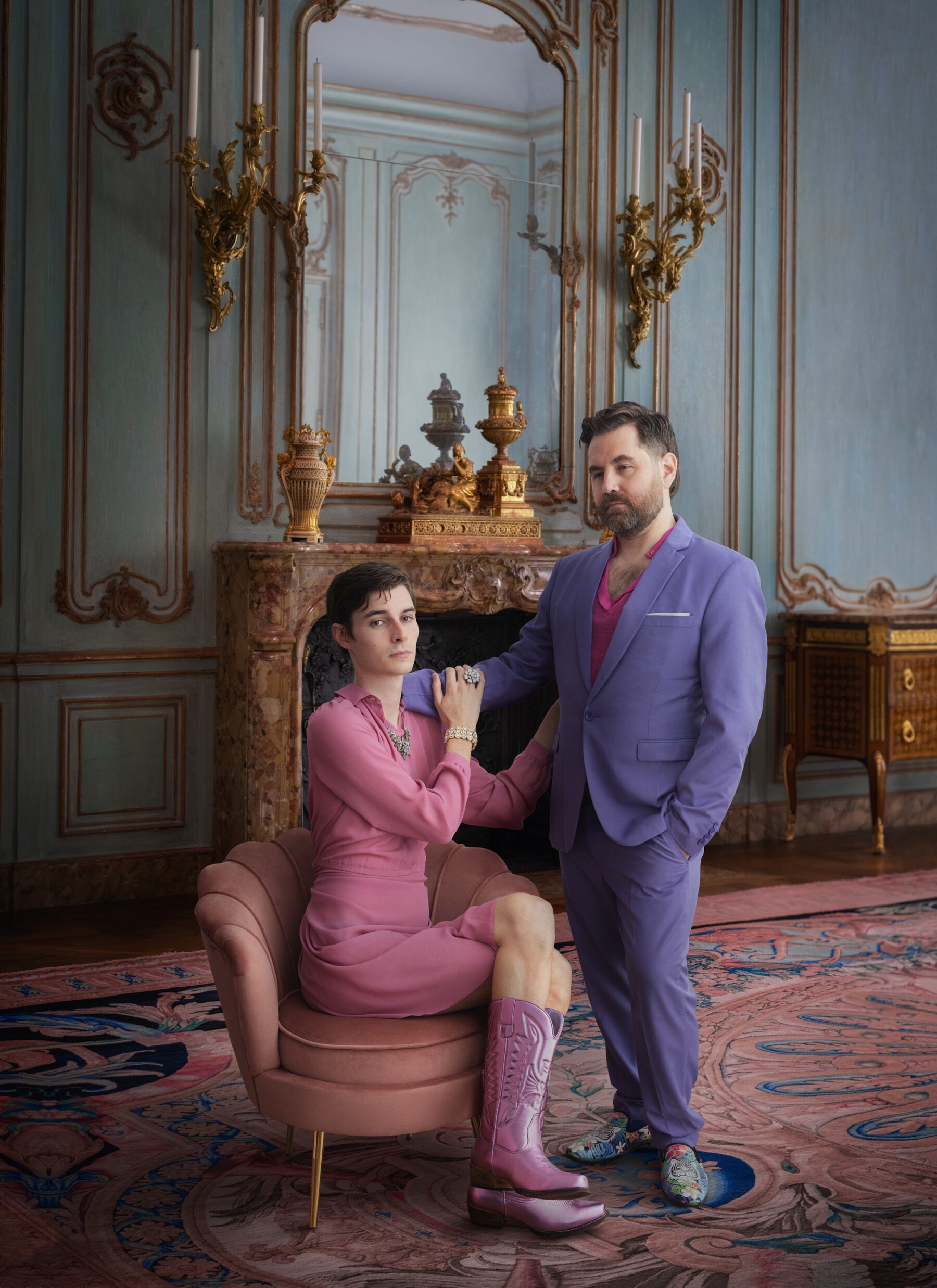 Two male-presenting figures are wearing pastel clothing, one is seated and they are touching, both figures are located in a room decorated in 18th century fashion