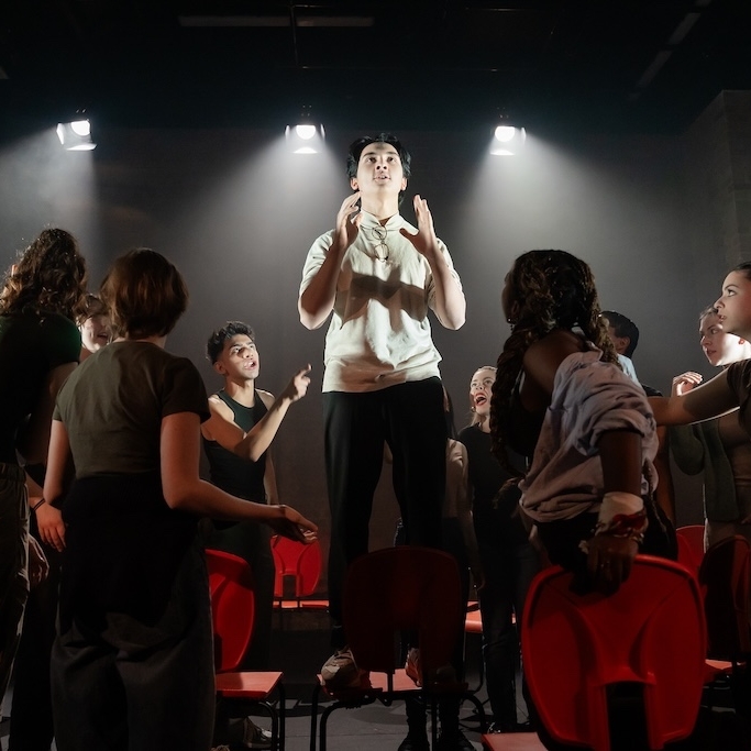 Action shot of the spring 2024 Shakespeare in Performance at RADA final presentation of Julius Caesar. An actor stands on a bright orange chair in mid-performance, lit by spotlights overhead, while other actors surround him on stage.