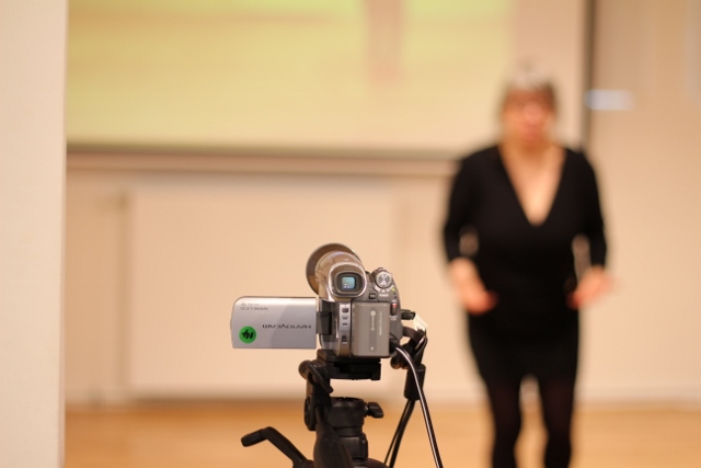 Video camera in the foreground with Sarah Thorn out of focus in the background.