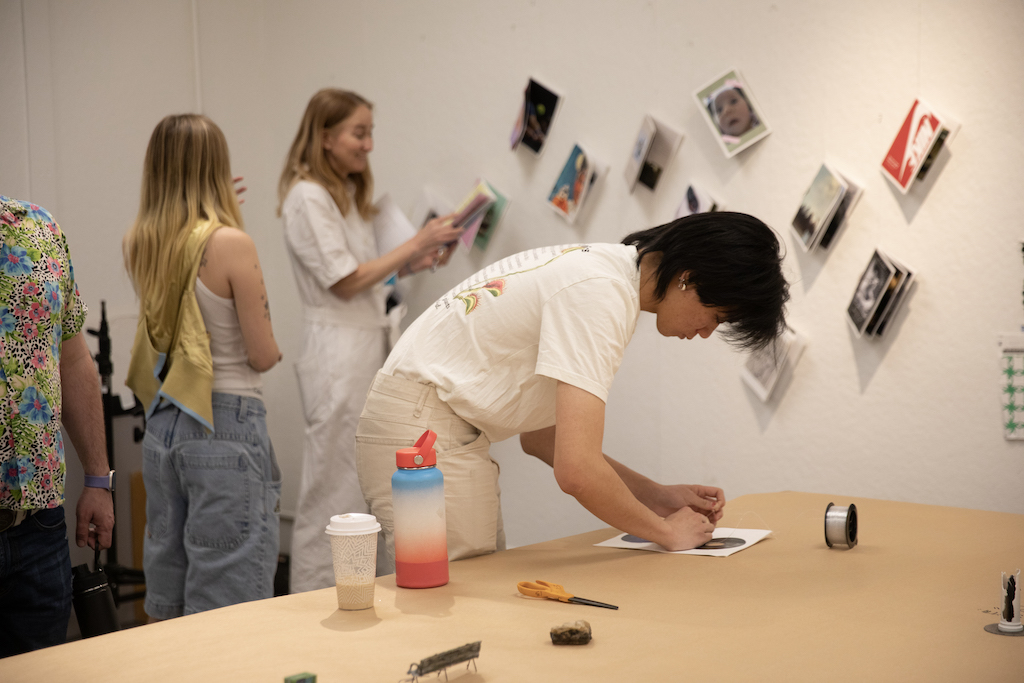 Photo leaning over a table, working on a photo while two students stand in the background looking at photograph booklets pinned to the wall