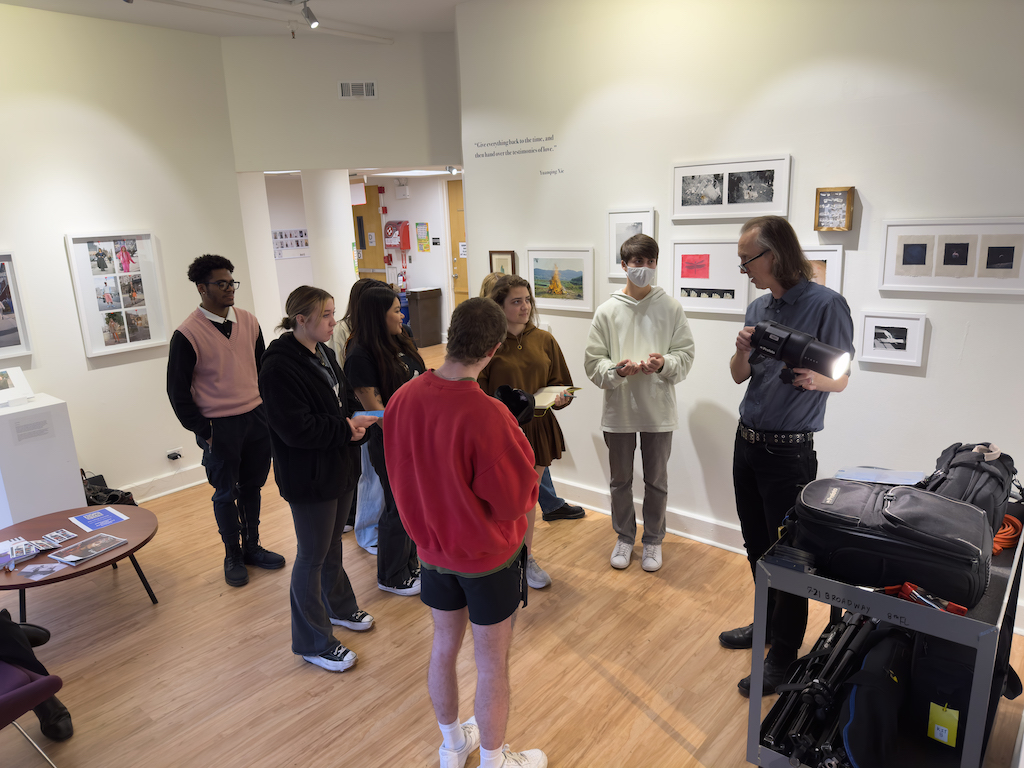 A group of students stand in the Department of Photography and Imaging listening to a professor who is demonstrating with a camera light